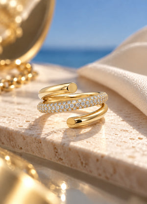 A pair of gold open wrap rings, one featuring a row of pavé-set clear stones, displayed on a light stone surface with a blurred beach background.