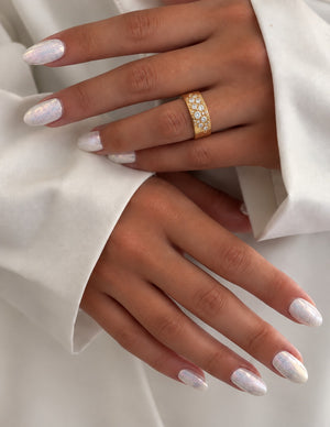 A close-up of a person's hands with white glitter nail polish, wearing a gold ring set with small clear stones on one finger, against a white fabric background.
