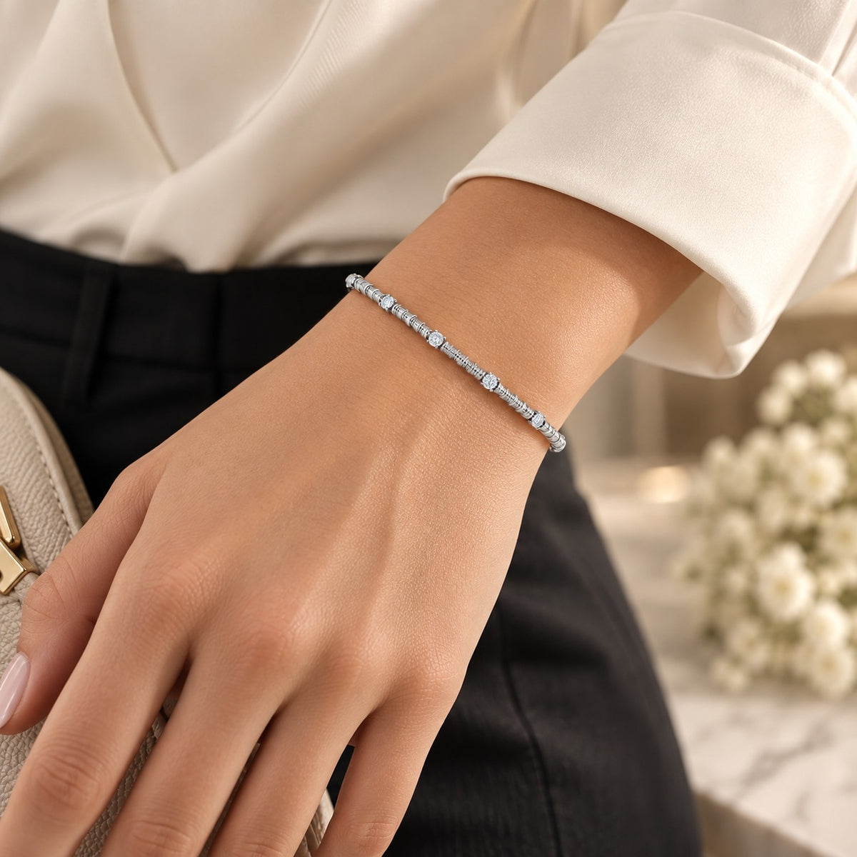 A delicate silver tennis bracelet with clear rectangular gemstones worn on a woman's wrist, paired with a cream blouse and black trousers.