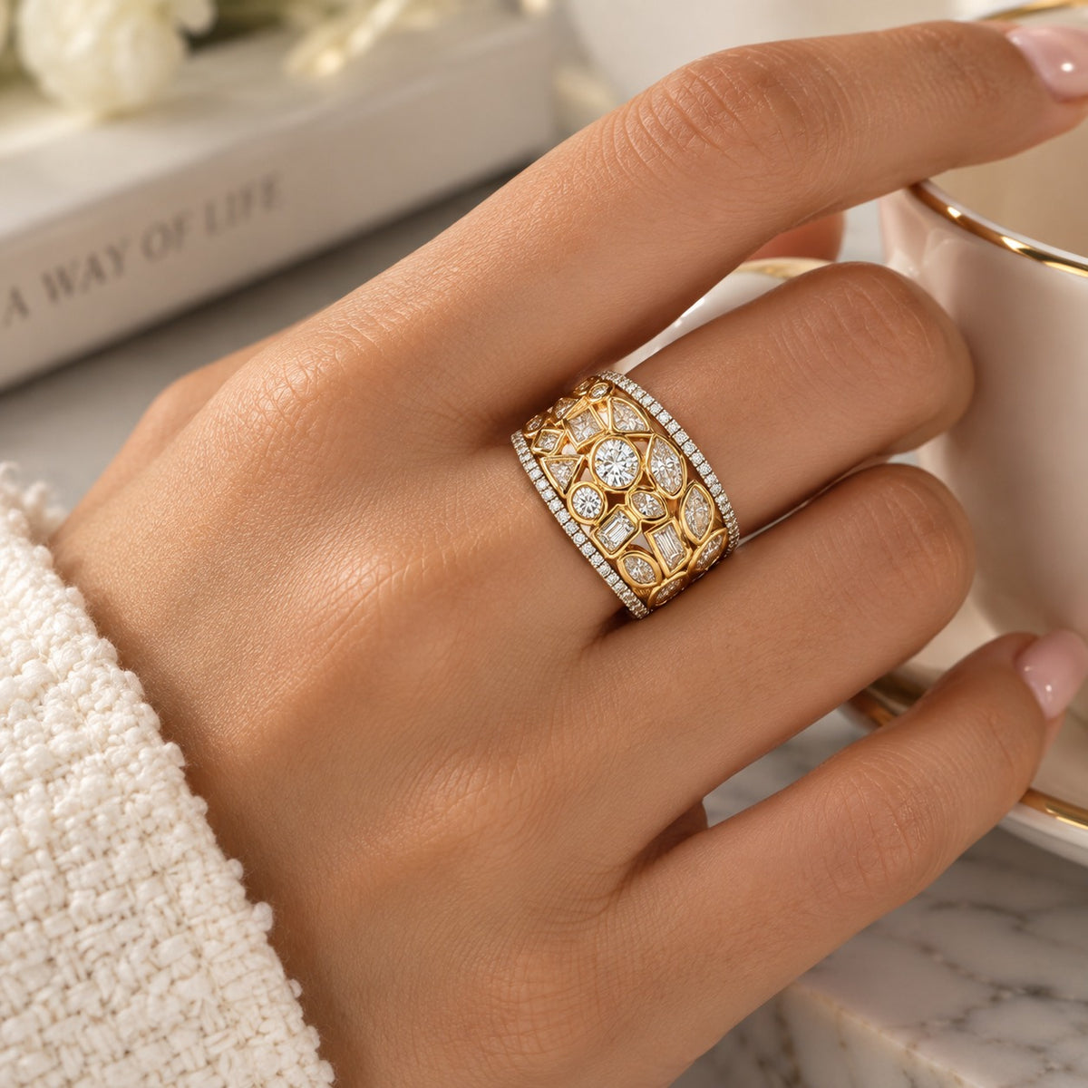 A close-up of a hand wearing a wide gold ring with pavé-set clear stones and an openwork floral pattern, styled with a cream knit sleeve and a white teacup on a marble surface.