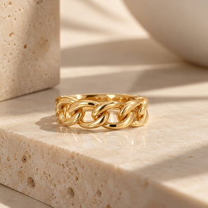 A polished gold chain-link ring resting on a beige travertine stone surface with warm, directional sunlight and soft shadows.