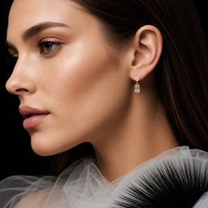 A close-up side profile of a woman wearing a delicate gold drop earring with a clear rectangular gemstone, set against a dark background.