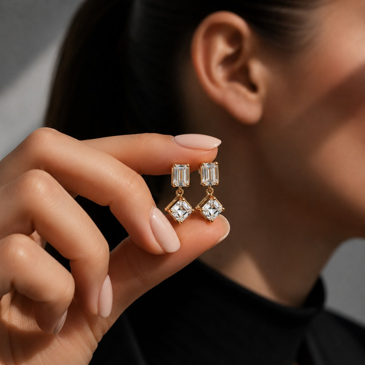 A close-up of a person's hand holding a pair of gold drop earrings with clear rectangular and diamond-cut gemstones, with the person's ear and neck visible in the background.