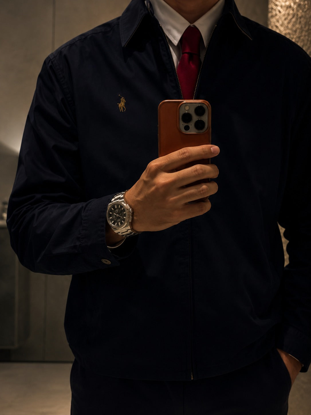 A man takes a mirror selfie showing a dark navy blue zip-up jacket with a small gold embroidered polo player on the left chest, worn over a white collared shirt and a red tie.