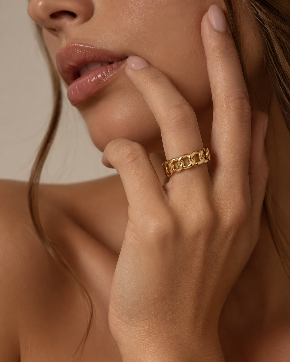 A close-up lifestyle shot of a model wearing a chunky gold chain-link ring on her finger, with her hand resting against her cheek.