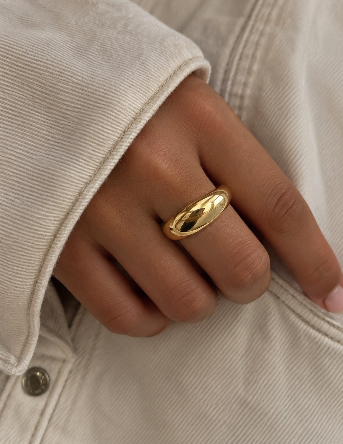 A close-up of a hand wearing a polished gold ring, set against a light beige denim jacket.