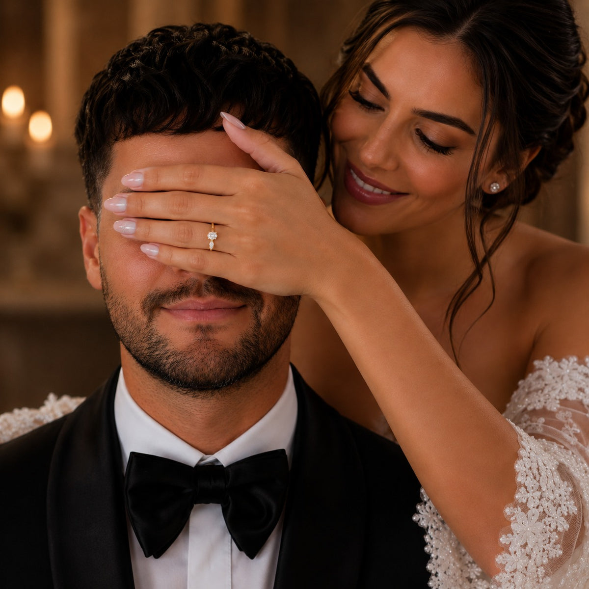 A smiling bride in a white lace wedding dress covers a man's eyes with her hand, which features a delicate diamond ring on the ring finger. He is wearing a black tuxedo with a bow tie, and the background is a softly blurred warm-toned indoor setting.