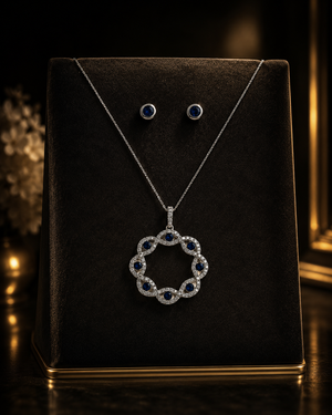 A Liria Jewelry set crafted in 14K white gold, featuring a pendant necklace and matching stud earrings accented with deep blue gemstones, elegantly displayed on a dark marble surface beside a black Liria Jewelry branded jewelry box.