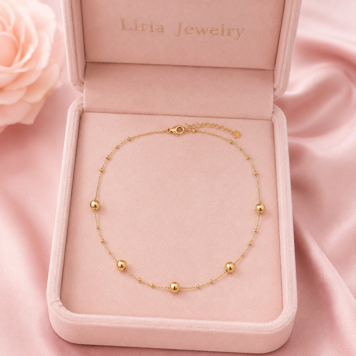 A delicate gold chain anklet with five small polished gold beads, displayed inside an open pink velvet jewelry box with 'Liria Jewelry' printed inside the lid, set against a pink satin background.