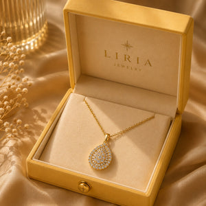 A gold teardrop pendant necklace with clear stones, displayed inside an open gold LIRIA JEWELRY box on a champagne-colored satin fabric background.