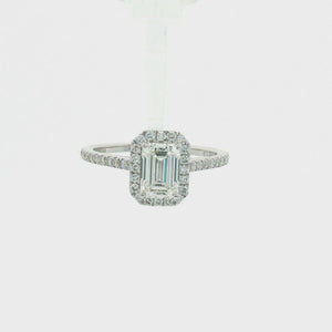 An emerald-cut halo ring featuring a central baguette diamond surrounded by a double halo of smaller round diamonds, with a pavé-set band in a silver-colored metal, shown on a plain white background.