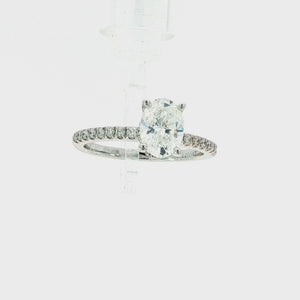 An oval diamond engagement ring in a silver-colored metal setting, featuring a halo of small diamonds and a pavé band with round diamonds, shown on a plain white background.