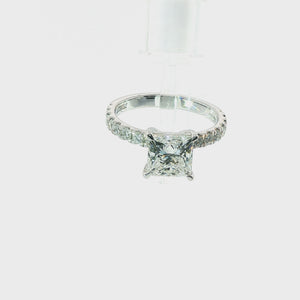 An engagement ring featuring a brilliant round-cut diamond center stone in a four-prong setting, with a pavé band set with small round diamonds, shown on a solid white background.