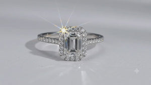 An emerald-cut halo ring featuring a central baguette diamond surrounded by a double halo of smaller round diamonds, with a pavé-set band in a silver-colored metal, shown on a plain white background.