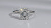 An emerald-cut halo ring featuring a central baguette diamond surrounded by a double halo of smaller round diamonds, with a pavé-set band in a silver-colored metal, shown on a plain white background.
