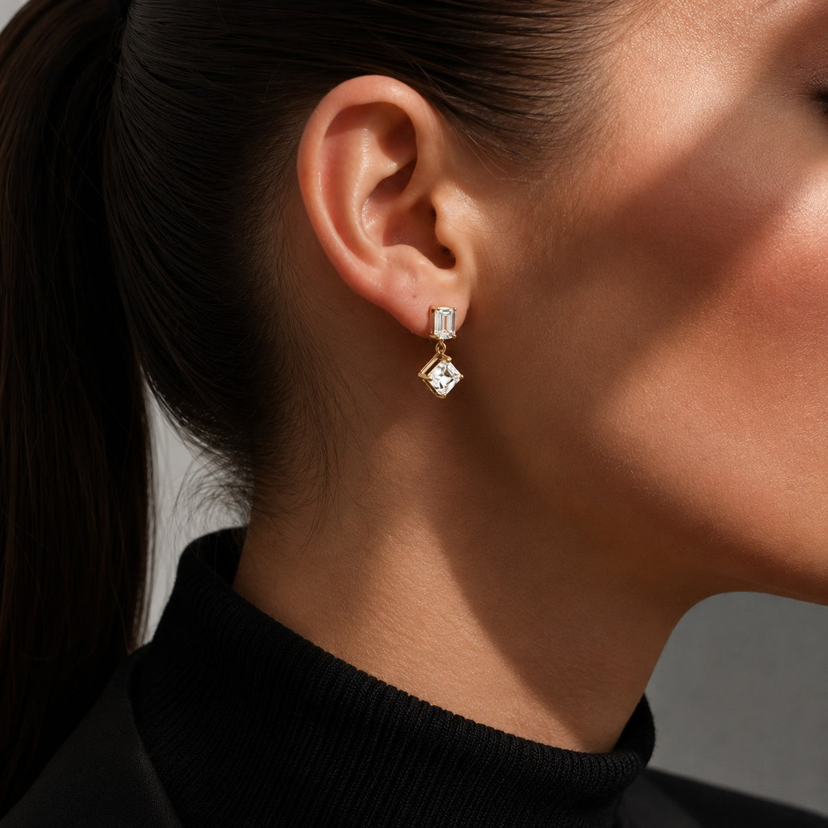 A close-up of a model's ear wearing a gold drop earring with a rectangular clear gemstone and a dangling clear gemstone in a diamond-cut setting.
